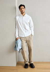 Witte overhemelf, beige broek, zwarte schoenen; houdt een lichtblauwe spijkerjack vast. Gladde stof, losse pasvorm, neutrale kleuren, eenvoudig ontwerp.