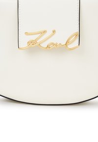 Balta ādas soma ar zelta metāla slīpo "Karl" logo detaļu apakšcentrā un melnu apmalu gar malu.