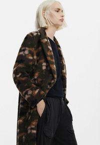 Brun og grøn camouflage uldfrakke med notched revers, oversized silhuet og sidelommer, parret med en sort top og mørke bukser.