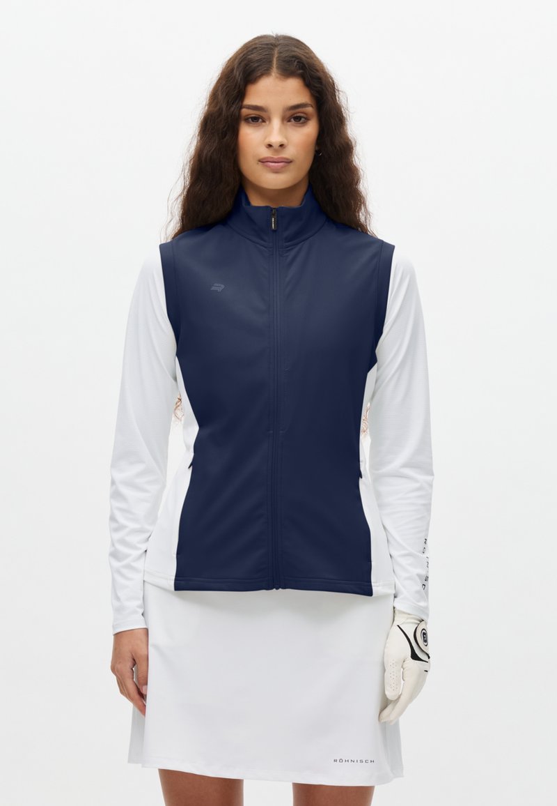 Vrouw draagt een marineblauw mouwloos vest over een wit shirt met lange mouwen en een witte rok, met een witte golfhandschoen aan haar rechterhand.