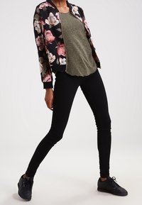 Floral Bomberjacke in Schwarz mit rosa und cremefarbenen Rosen, grünes T-Shirt, schwarze Skinny-Jeans und schwarze Sneaker mit strukturierten Sohlen.