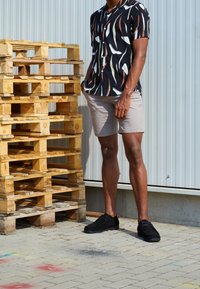 Zwart shirt met korte mouwen met witte en oranje patronen, beige korte broek en zwarte schoenen, staand naast gestapelde houten pallets op het trottoir.