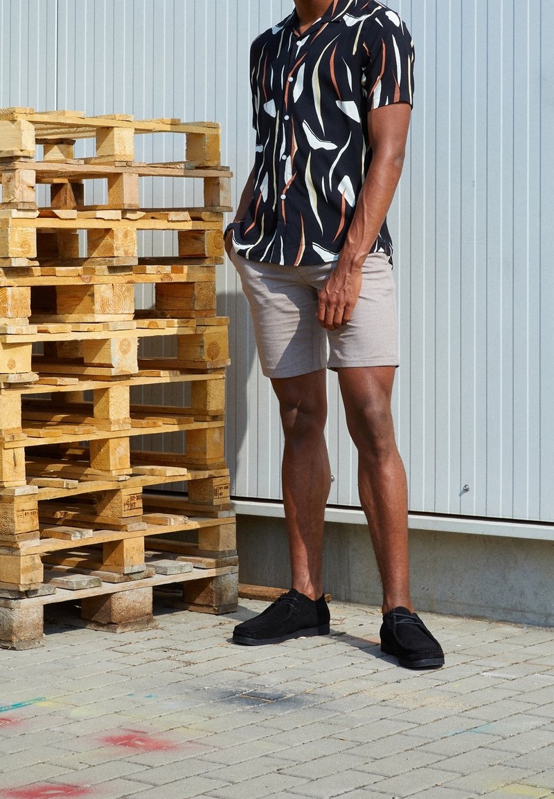 Zwart shirt met korte mouwen met witte en oranje patronen, beige korte broek en zwarte schoenen, staand naast gestapelde houten pallets op het trottoir.