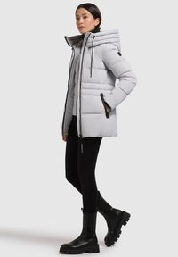 Lys grå dunjakke med hætte, lodret quiltet design, sidelommer med sorte lynlåse, båret over sorte leggings og kraftige sorte støvler.