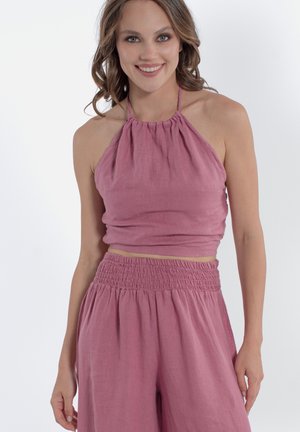 Giovane donna sorridente, indossa un top corto rosa senza maniche con scollo halter e pantaloni abbinati a vita alta e gamba larga su sfondo semplice.