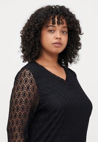 Μαύρη μπλούζα με δαντελένια μανίκια με κυκλικά σχέδια, σχέδιο με V-ντεκολτέ και υφή υφάσματος, επιδεικνύοντας εφαρμοστό σιλουέτα.