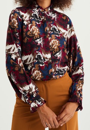Frau trägt eine langärmelige Blumenbluse in Burgunder, Blau, Braun und Weiß kombiniert mit hoch taillierten Senfhosen.