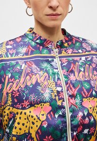 Blouson aviateur coloré avec un motif floral et léopard. Fermeture zippée, encolure ronde, et tons vibrants de violet, jaune et vert.