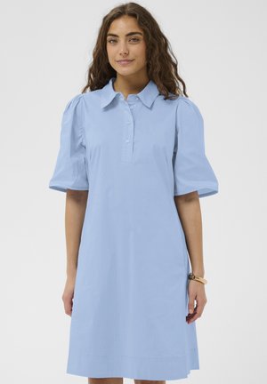 Femme aux longs cheveux bouclés portant une robe bleu clair à manches courtes boutonnée, debout devant un fond uni.