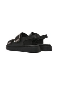 Sandalias de cuero negro con correas ajustables, que presentan detalles en metal plateado, una suela plana y un diseño de espalda abierta.