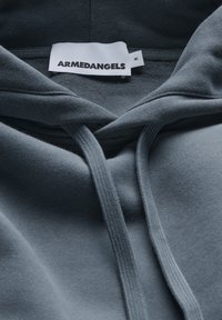 Grauer Kapuzenpullover mit weicher Textur, ausgestattet mit einer Kängurutasche und Kordelzügen. Das Etikett trägt die Aufschrift "ARMEDANGELS". Größenetikett enthalten.
