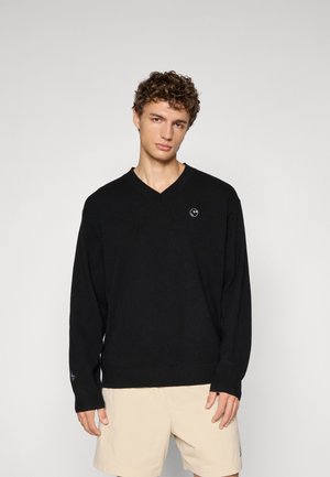 Svart V-hals genser laget av et mykt materiale, med en liten logo på brystet og ribbestrikkede mansjetter. Kombinert med beige shorts.