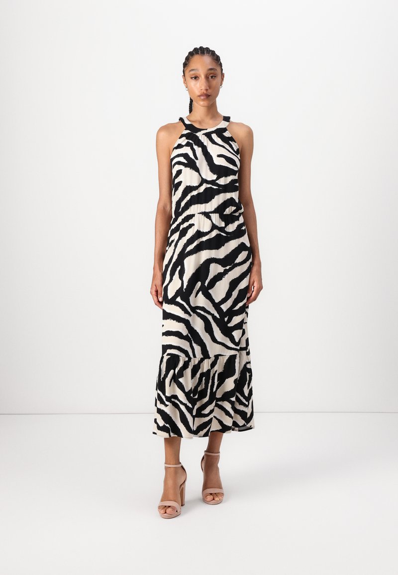 Robe maxi avec un motif zèbre, présentant un encolure halter, des couleurs noir et blanc, et un ourlet volanté. Portée avec des sandales à talons nus.
