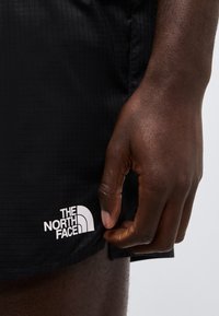 Tumedanahaline käsi reguleerimas musti lühikesi pükse, mille ääres on The North Face'i logo, ühevärvilise tausta ees.