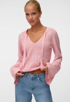 Jeune femme aux cheveux plaqués en arrière porte un pull en maille rose à larges manches et un jean bleu clair, posant devant un fond blanc.