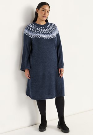 Dunkelblaues Strickkleid mit einem gemusterten Einsatz in Hellblau, lockeren langen Ärmeln und einer lässigen Passform, kombiniert mit schwarzen Leggings und Stiefeln.