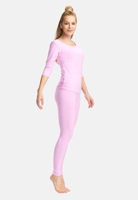 Blond naine, kes tasakaalustab varvastel, kandes heleroosat pika varrukaga pusa ja sobivaid retuuse, seisab valge tausta ees.