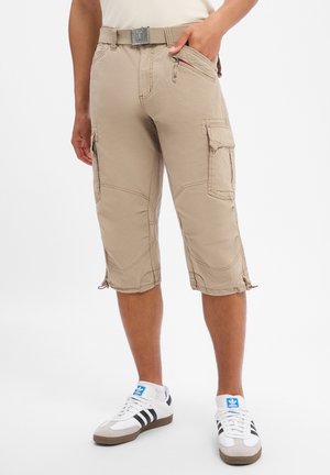 Person wearing beige cargo shorts with multiple pockets and a belt, paired with white Adidas sneakers with black stripes.