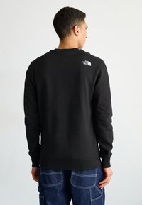 Sudadera negra de manga larga, con puños y dobladillo de canalé, que presenta un pequeño logo blanco en la parte superior de la espalda. El material parece suave y grueso.