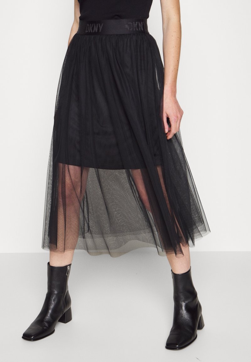 Femme portant une jupe midi en maille noire à couches avec une ceinture à logo DKNY et des boots noires à talons blocs.