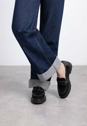 Zapatos slip-on de ante negro con un detalle de cadena texturizada, combinados con jeans de mezclilla azul que tienen dobladillos grises enrollados y costuras visibles.