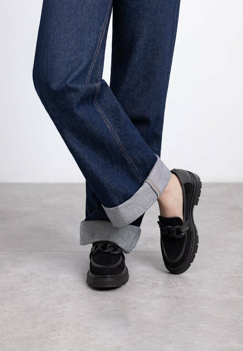 Zapatos slip-on de ante negro con un detalle de cadena texturizada, combinados con jeans de mezclilla azul que tienen dobladillos grises enrollados y costuras visibles.