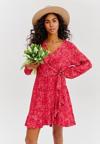 Červené zavinovací šaty s dlouhými rukávy, páskem na zavazování v pase a volánkovým lemem. Květnatý vzor, nošené s širokým kloboukem v světlé barvě a držením tulipánů.