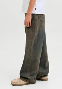 Široke maslinasto zelene traperice s izblijedjelom teksturom i stražnjim džepovima, u kombinaciji s bež cipelama na klizanje.