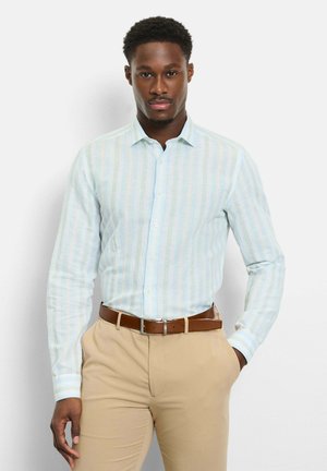 Mann trägt hellblau-weiß gestreiftes Hemd, in beige Hose mit braunem Gürtel gesteckt, steht vor weißem Hintergrund.