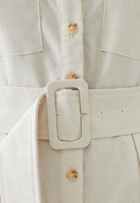 Valkoinen pellavamekko, jossa on napitus, suuret rintataskut ja leveä vyö suoralla soljella, jossa on useita reikiä.