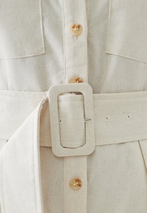 Leinenkleid in hellem Beige mit Knopfverschluss, großen Brusttaschen und einem breiten Gürtel mit rechteckiger Schnalle und mehreren Löchern.