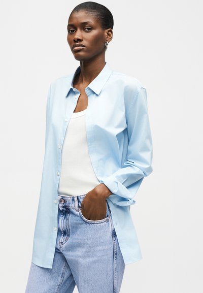 Γυναίκα με κοντά μαλλιά που φοράει ανοιχτό μπλε πουκάμισο με κουμπιά πάνω από λευκή φανέλα και ανοιχτό μπλε τζιν, το χέρι της στην τσέπη, με ουδέτερη έκφραση.
