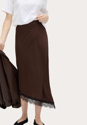 Person in einem weißen Hemd und einem dunkelbraunen Midirock mit Spitzenbesatz, hält eine dazu passende dunkelbraune Jacke in einer Hand.