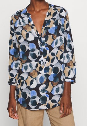 Bluzka z długim rękawem na guziki z abstrakcyjnymi okrągłymi wzorami w kolorach niebieskim, beżowym, czarnym i białym, noszona z beżowymi spodniami.