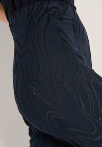 Close-up van donkerblauwe broek met abstracte zwarte gestikte patronen en een zichtbare achterzak.