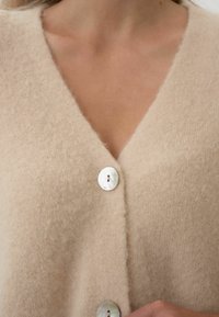 Beżowy sweter z dzianiny z dekoltem w serek, z dwoma okrągłymi guzikami z masy perłowej i miękkim, teksturowanym materiałem.
