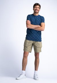 Chemise bleu marine à manches courtes avec motifs verts, associée à un short cargo beige. Des baskets blanches et des chaussettes montantes complètent la tenue.