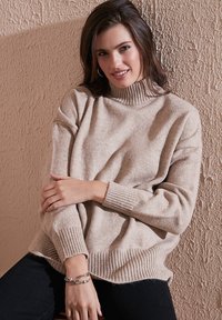 Beež turtleneck kampsun ribitud varrukate ja äärtega, valmistatud tekstureeritud kangast. Mudel poseerib istudes tekstureeritud seina vastu, kandes musti teksaseid.