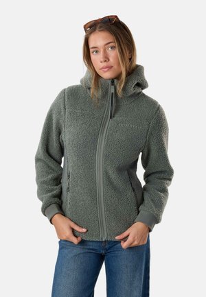 Frau trägt graue Fleecjacke mit Reißverschluss und Kapuze sowie Sonnenbrille auf dem Kopf, Hände ruhen nahe der Taille, steht vor einfarbigem Hintergrund.