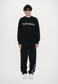 Svart hettegenser og bukser med "No Problemo" trykt i hvitt. Solbriller og hvite sneakers fullfører antrekket.