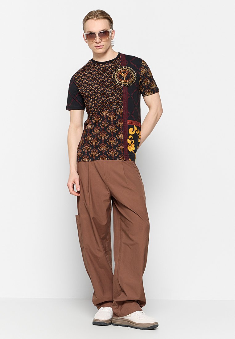 Schwarzes T-Shirt mit kurzen Ärmeln und komplizierten goldenen und roten Mustern, kombiniert mit lockeren braunen Hosen und weißen Sneakers. Accessoires sind Sonnenbrillen.