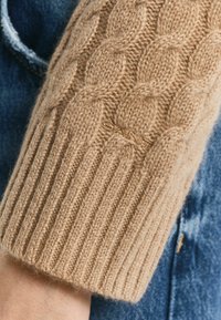 Beiger, kabelgestrickter Pullover mit einem gerippten Bündchen an den Ärmeln. Die weiche Textur hebt sich von einem blauen Denim-Hintergrund ab. Sichtbare, detaillierte Nähte.