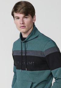 Sudadera con capucha en color azul verdoso con paneles a rayas en negro y gris. Cuenta con un bolsillo tipo canguro, cordón ajustable y detalles de logo en relieve.