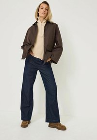 Hnědá lehká bunda přes krémový pletený svetr, spárovaná s tmavě modrými kalhotami širokého střihu a hnědými semišovými botami. Jednoduché pozadí.