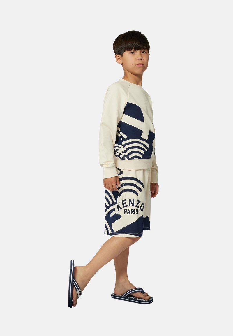 Lichtbeige sweatshirt met blauwe grafische ontwerpen, gecombineerd met blauwe geblokte shorts. De outfit wordt aangevuld met marineblauwe flip-flops.