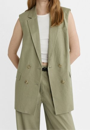 Kobieta w bezrękawnym oliwkowozielonym dwurzędowym marynarce i dopasowanych spodniach, nosząca biały prążkowany top na brzuch.