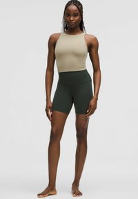 Žena stojaca naboso, oblečená v béžovom bezrukávovom crop top a tmavozelených dlhých cyklistických šortkách, na jednofarebnom pozadí.