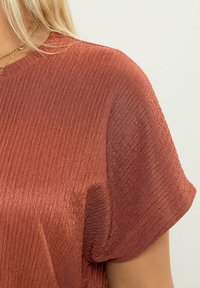 Rustinvärinen paita, jossa on teksturoitu, ribbikangas. Lyhyet hihat ja pyöreä pääntie korostavat sen sulavaa muotoilua ja modernia vetovoimaa.