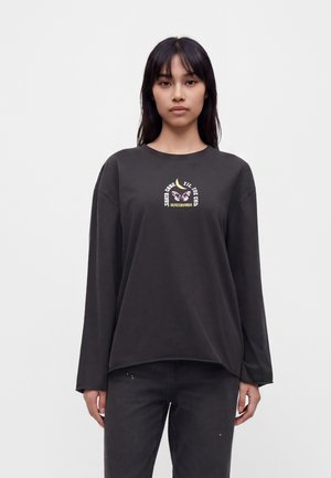 Femme aux longs cheveux foncés portant un t-shirt noir à manches longues avec un petit motif coloré et un texte sur la poitrine, debout devant un fond uni.