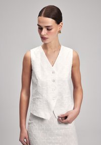 Witte zonder mouwen vest met een bloemig reliëfpatroon, v-halslijn en vijf knopen. Beschikt over twee voorzakken en een gestructureerde pasvorm.
